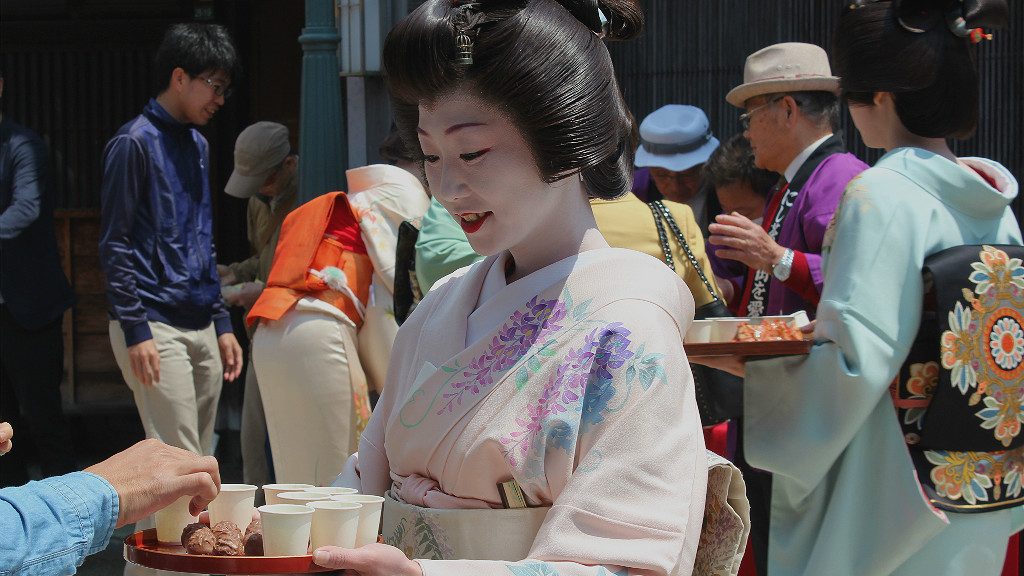 a geisha in a wisteria-themed kimono serves sake on the street