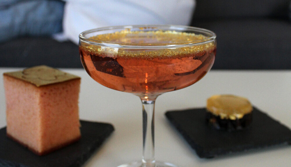 Pink champagne with gold flakes floating on top. In the background, gold topped sakura-flavored castella cake and a gold and red sweet bean Japanese sweet.