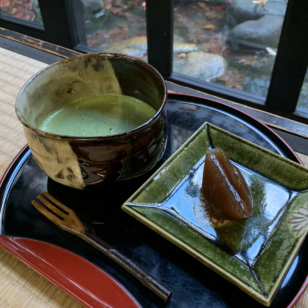 a bowl of matcha green tea and a wagashi Japanese bean paste sweet served on a tatami mat in front of a Japanese garden